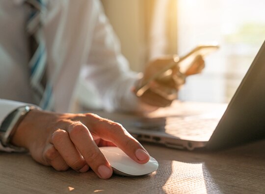 mão esquerda mexendo no computador e utilizando o mouse enquanto a mão direita segura um celular