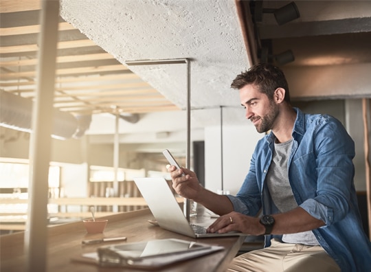 home de camiza jeans utilizando o celular  em uma mesa com papeis e notebook a sua frente.