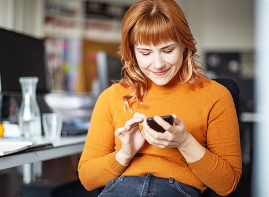 cliente Itaú usando uma blusa laranja em frente a uma mesa com uma tela de computador utilizando seu celular