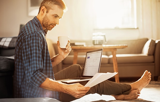 Cliente Itaú sentado no chão com um notebook no colo, tomando um café enquanto mexe com papelada.
