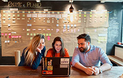 três colaboradores do departamento varejo sentados em frente a um notebook em uma sala de reunião, com vários post its colados na parede.