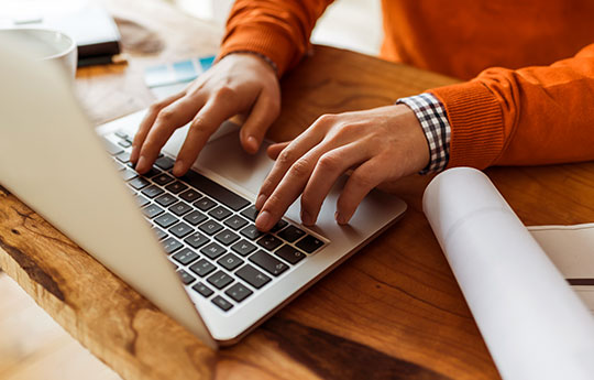 Pessoa com suéter laranja digitando no seu notebook em uma mesa de madeira