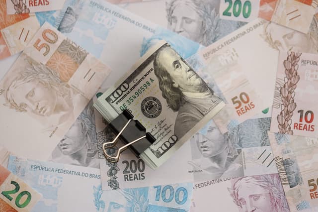 Maço de notas de dólar preso por um clipe sobre cédulas de real espalhadas, representando câmbio, investimentos e mercado financeiro.