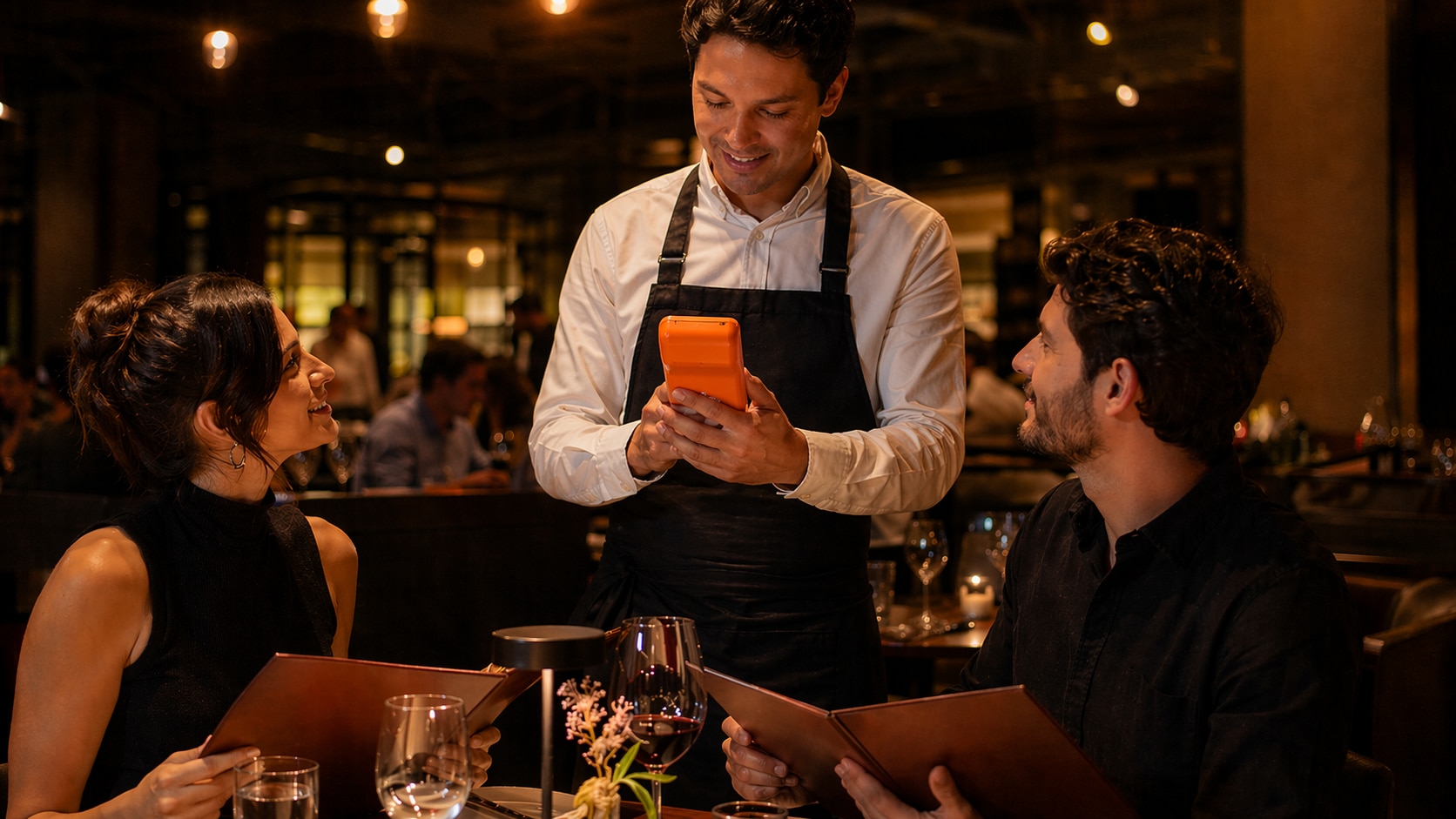 Cliente em um restaurante com iluminação baixa observa atendente em pé ao lado da mesa, utilizando um dispositivo móvel para registrar o pedido, enquanto duas pessoas sentadas seguram cardápios abertos sobre a mesa com taças e utensílios visíveis ao redor.
