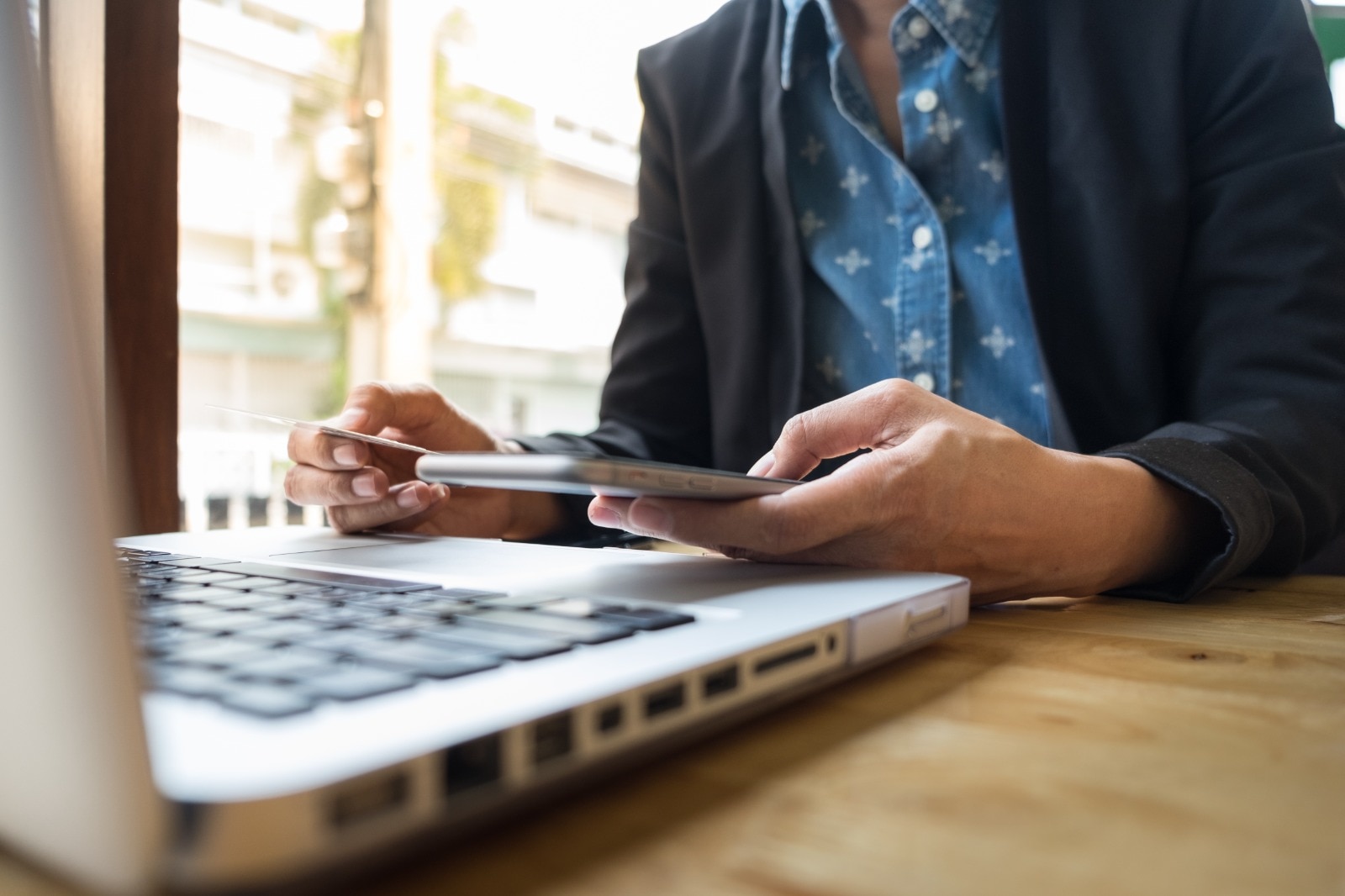Pessoa utilizando um smartphone enquanto segura um cartão bancário, ao lado de um notebook sobre uma mesa de madeira. A imagem remete à realização de operações financeiras digitais, pagamentos ou gestão financeira online.