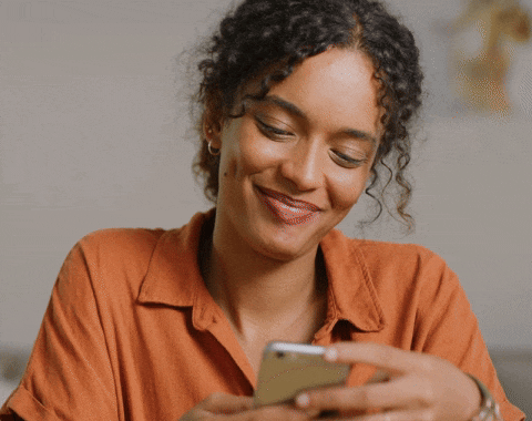 Mulher com camisa laranja sorrindo enquanto olha para o celular.
