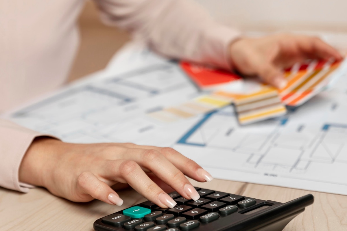 Mãos utilizando uma calculadora sobre uma mesa com documentos, gráficos e amostras de cores ao fundo, representando controle financeiro, emissão de documentos fiscais e gestão administrativa de um negócio.