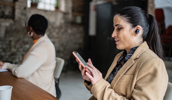 Mulher de fone de ouvido sentada em frente a uma mesa, prestando atenção no celular que está em sua mão.