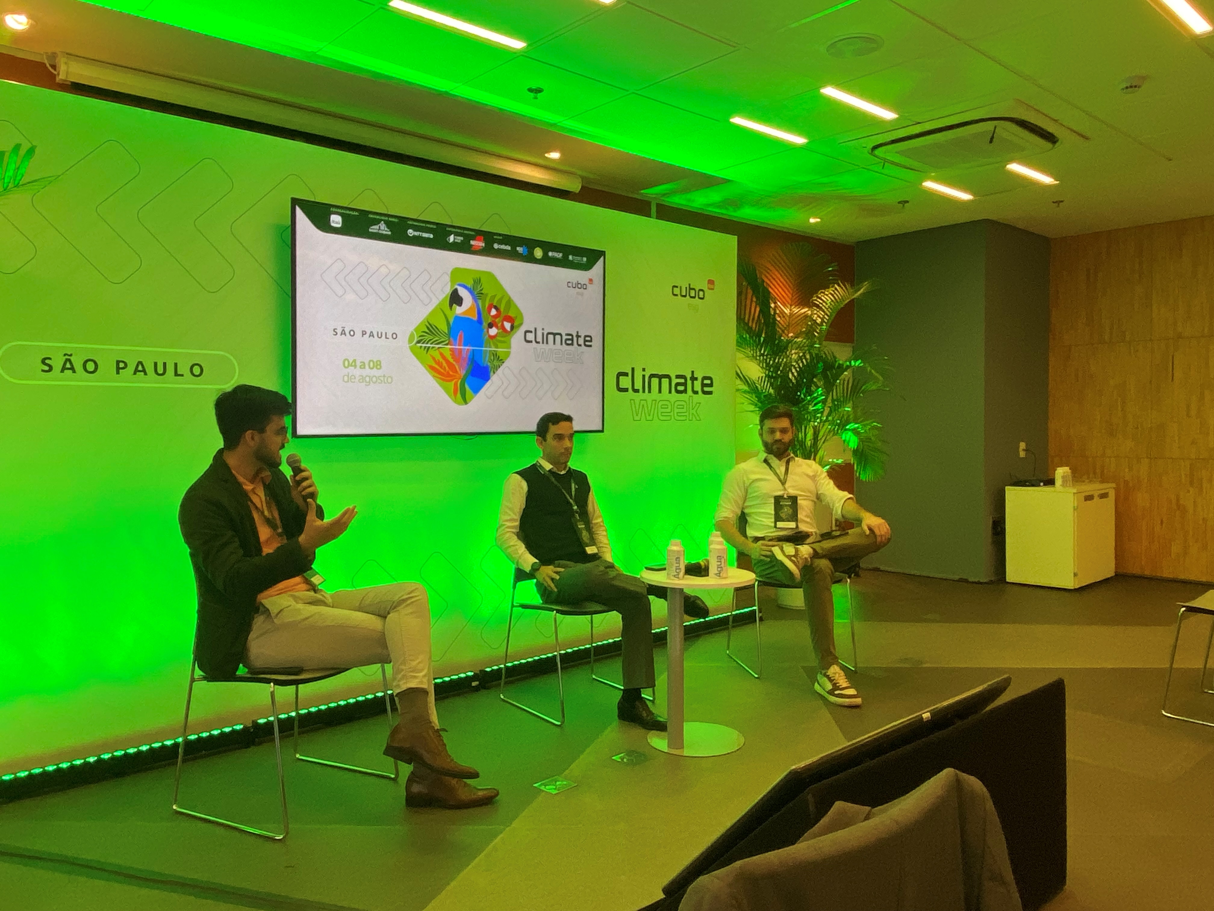 Os três palestrantes do painel, sentados em cadeiras. Ao fundo, telão verde com informações sobre o painel e o evento.