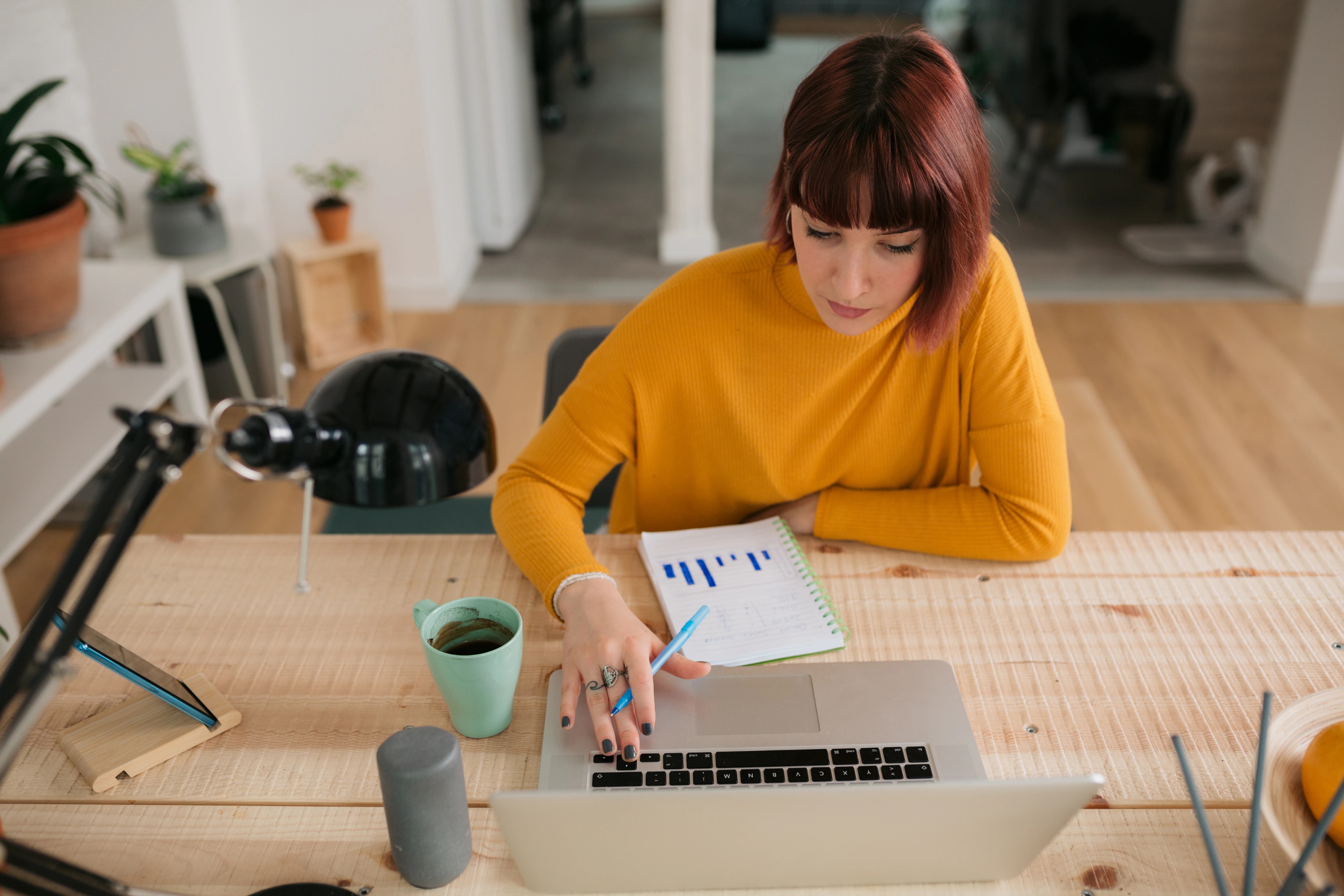 Na imagem há uma moça com cabelos lisos ruivos, ela veste uma blusa laranja e está sentada em uma mesa com um lápis na mão em frente a um notebook
