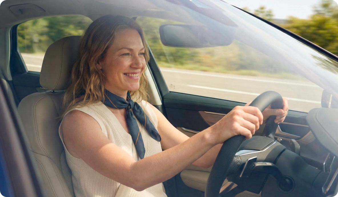 Cliente Itaú Personnalité dirigindo carro em estrada, com as mãos no volante e cinto de segurança. Interior claro do veículo e, ao fundo, paisagem com vegetação e pista sob luz natural entrando pela janela.