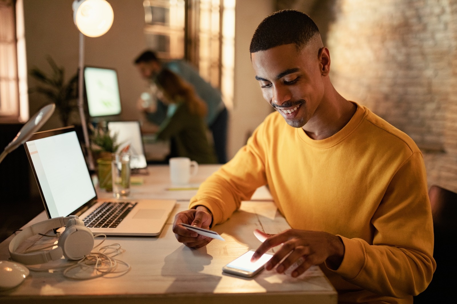 Pessoa sentada em uma mesa de trabalho utilizando um smartphone enquanto segura um cartão bancário. Sobre a mesa há um notebook aberto, fones de ouvido, documentos e um copo com água. Ao fundo, aparecem estações de trabalho e outras pessoas desfocadas em um ambiente corporativo com iluminação quente e paredes de tijolos aparentes.