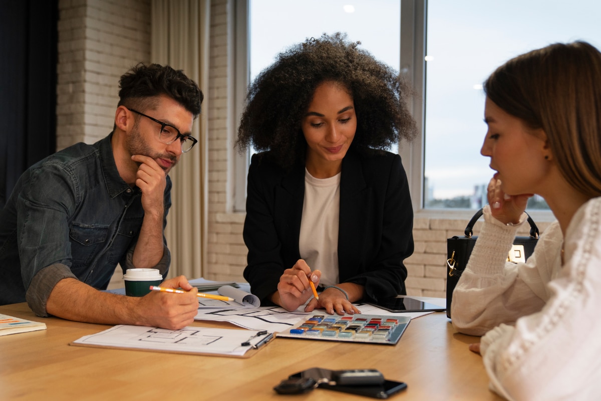 Três pessoas reunidas em uma mesa de trabalho analisando documentos, amostras de cores e anotações. Uma pessoa segura um lápis sobre uma cartela de cores enquanto as outras observam os materiais espalhados sobre a mesa. Há um copo descartável, uma prancheta, um tablet e um smartphone no ambiente. Ao fundo, vê-se uma janela ampla e paredes de tijolos claros em um espaço corporativo.