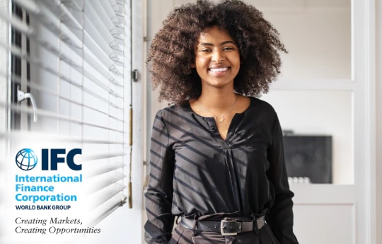 Mulher sorrindo em um escritório, com o logo do IFC (International Finance Corporation) no canto inferior esquerdo da imagem.