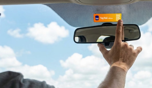 Mão posiciona tag veicular no para-brisa, próximo ao espelho retrovisor interno.