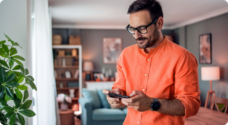 Um rapaz de óculos e camisa social laranja olha para a tela do seu smartphone e sorri despretenciosamente.
