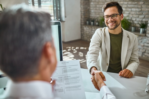 Duas pessoas apertam as mãos durante uma entrevista de emprego em um escritório. Em primeiro plano, uma pessoa segura um currículo enquanto a outra está sentada do outro lado da mesa, próxima a um computador.