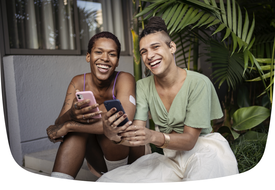 2 pessoas estão sentadas no quintal de casa, sorrindo e utilizando o celular.