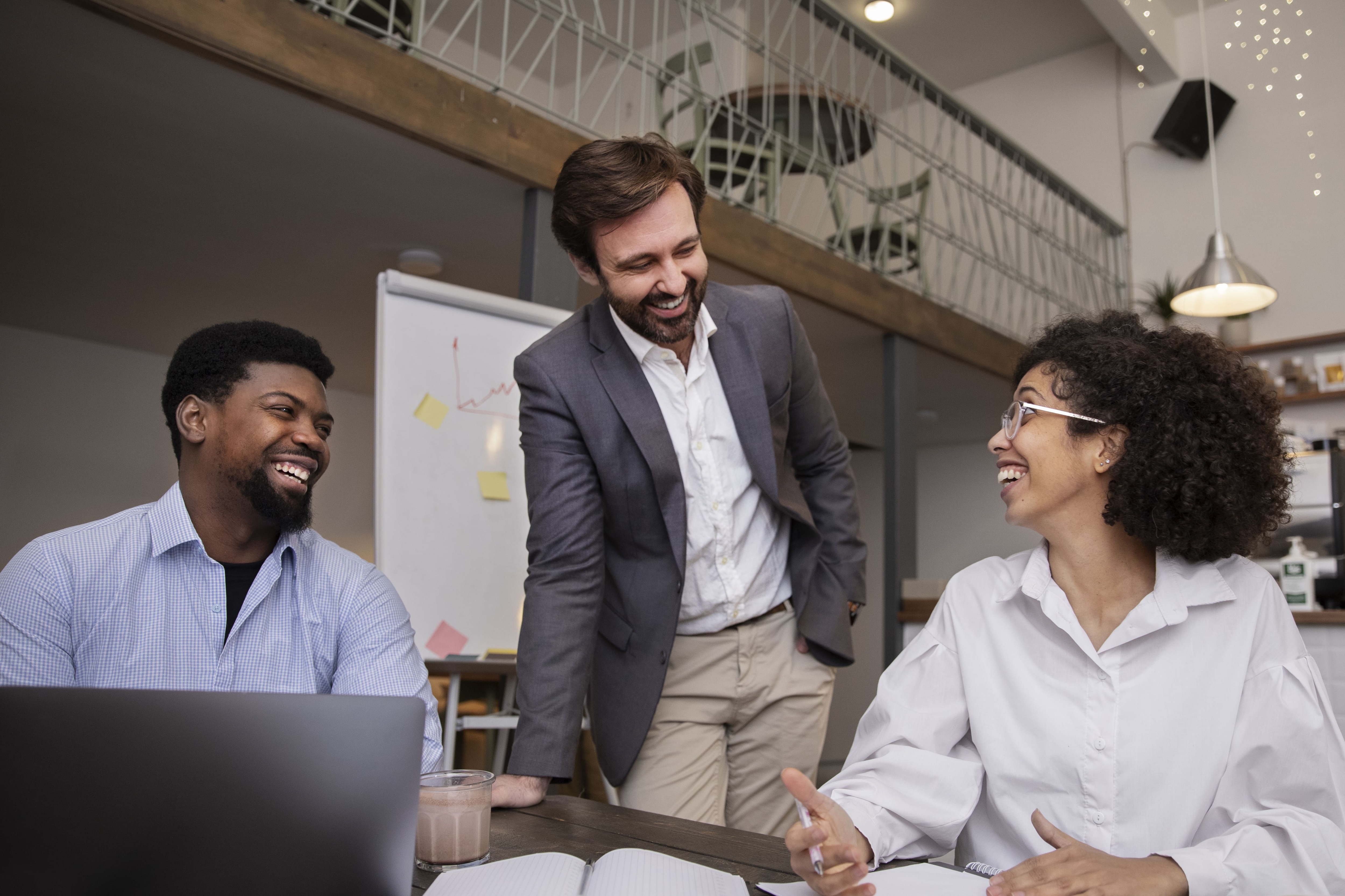 Equipe reunida em uma sala de reunião corporativa, com três pessoas ao redor de uma mesa de trabalho. Há um notebook aberto, cadernos de anotações e um quadro branco ao fundo, enquanto os participantes conversam durante uma reunião de equipe.