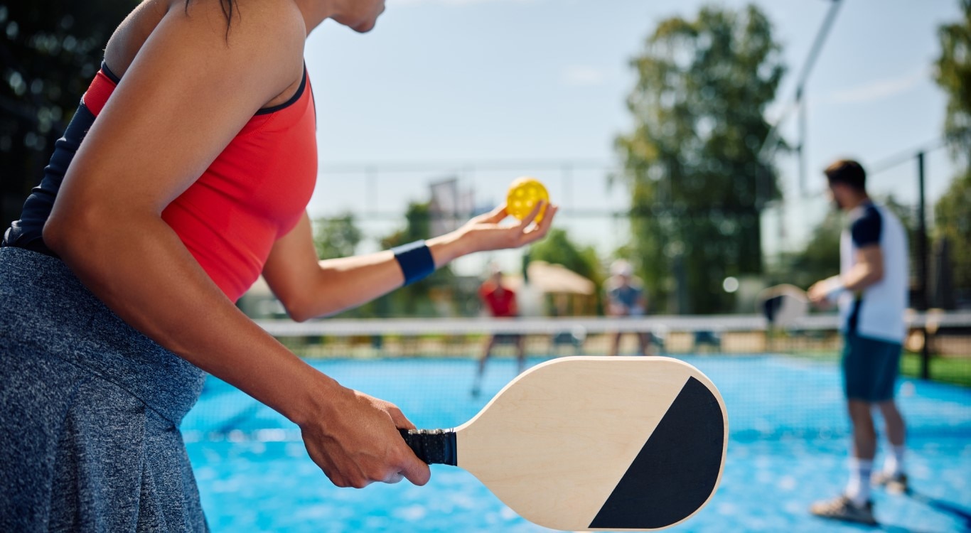 Uma pessoa vista de costas prepara-se para um jogo de pickleball, segurando uma bola amarela com uma mão e a raquete na outra. A pessoa está usando uma camiseta sem mangas vermelha e uma saia esportiva cinza. No fundo, outro jogador observa a cena, desfocado. O jogo está acontecendo em uma quadra azul com linhas brancas em um dia ensolarado.