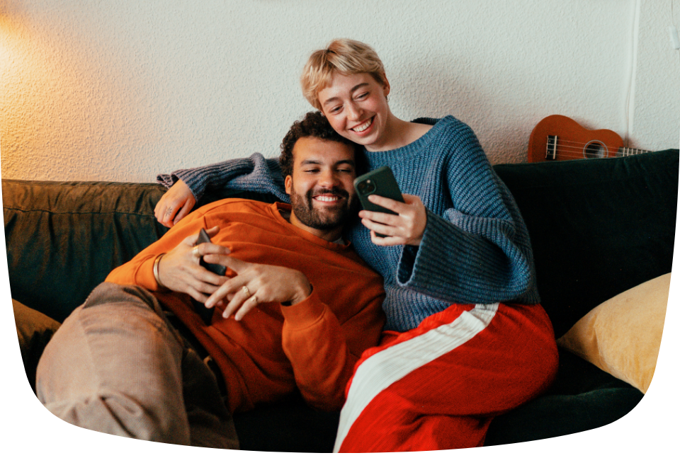Um homem e uma mulher está sentado no sofá. Os dois estão sorrindo. A mulher segura um celular e o homem está abraçado com ela.