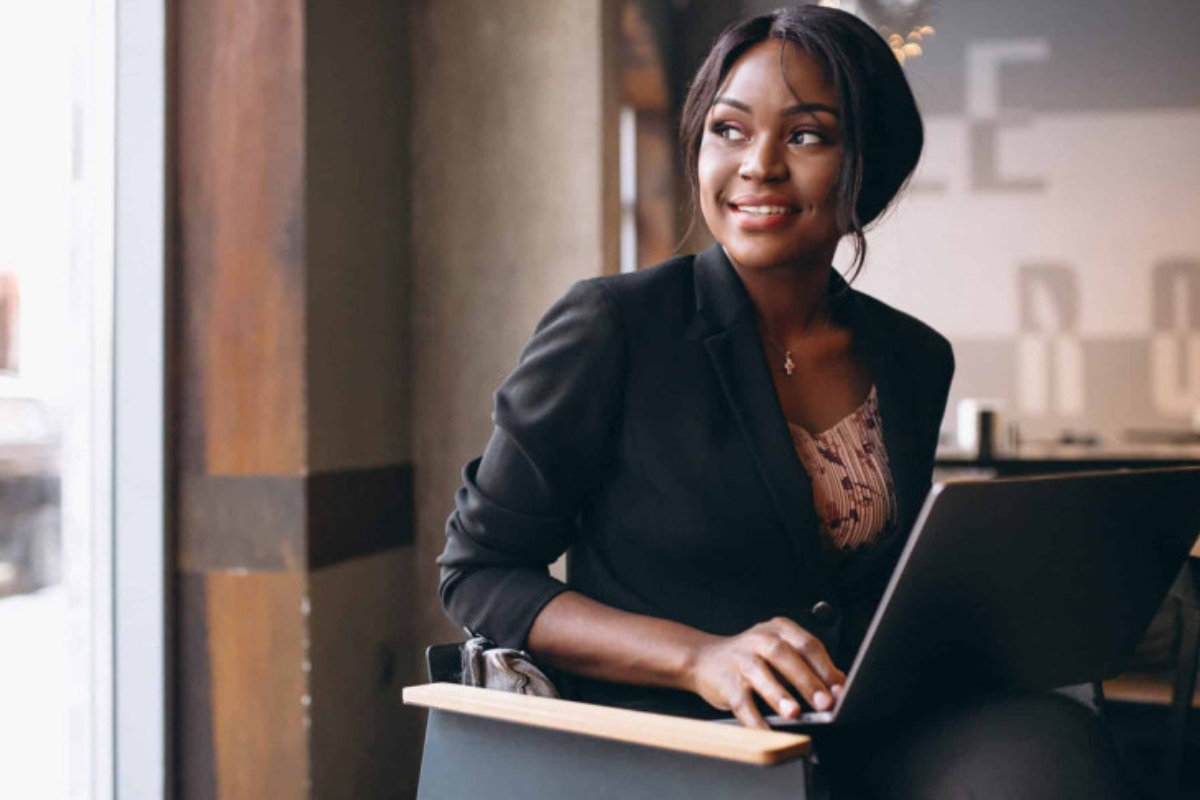Mulher sentada em ambiente profissional, sorrindo enquanto usa um notebook próximo a uma janela.