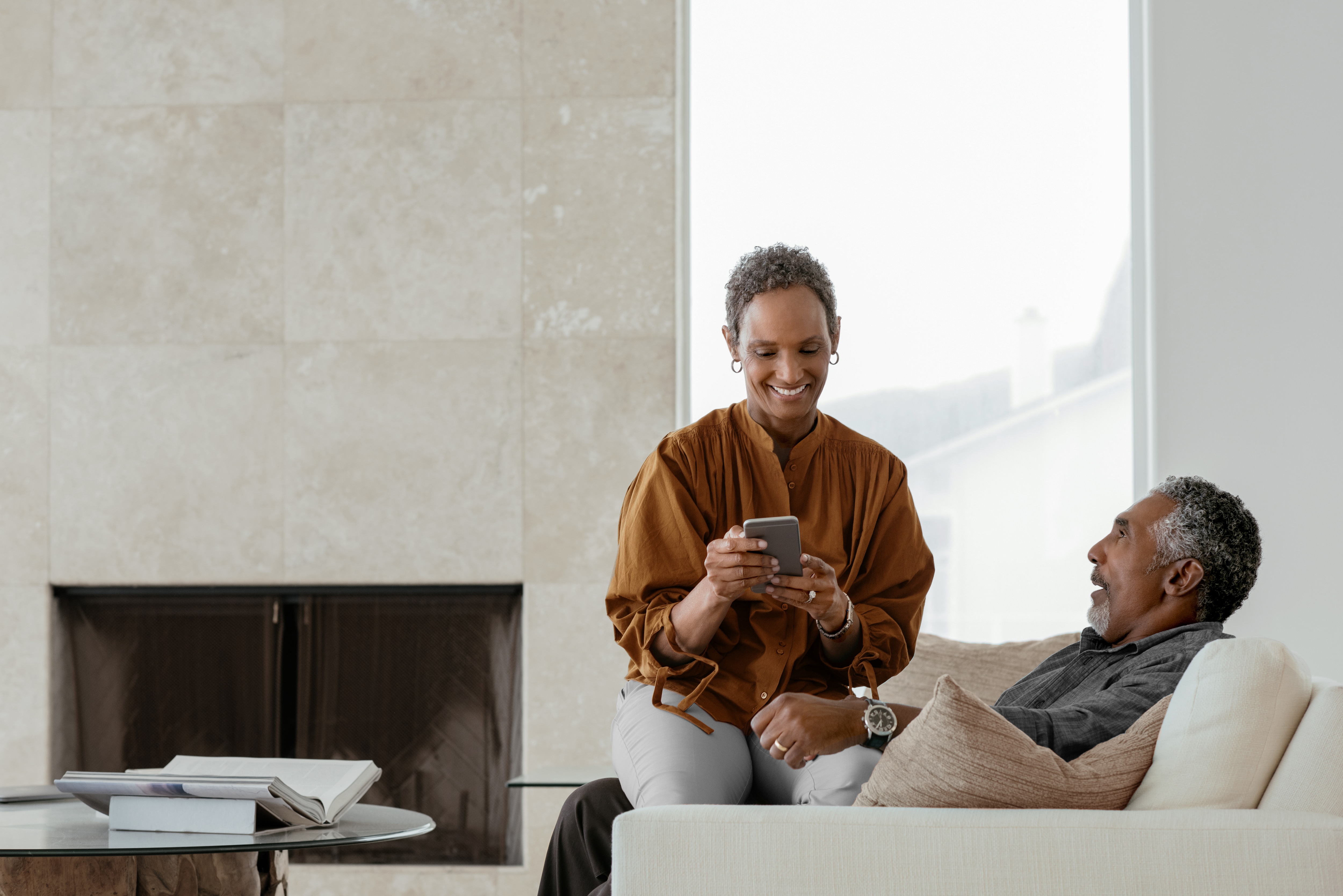 Homem e mulher clientes Itaú Private Bank conversam no sofá, ela sorri enquanto olha o celular.
