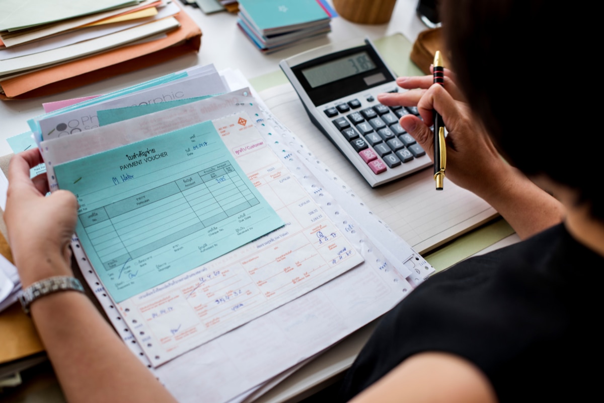 Pessoa analisando documentos financeiros e comprovantes impressos sobre uma mesa, enquanto utiliza uma calculadora e segura uma caneta para realizar cálculos e conferências de valores.