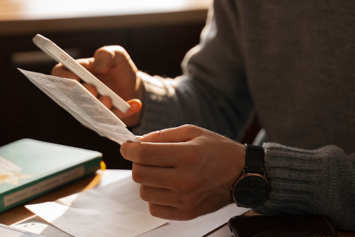 Pessoa segurando um comprovante em papel enquanto consulta informações no celular, sentada à mesa com documentos e um livro, em ambiente de trabalho