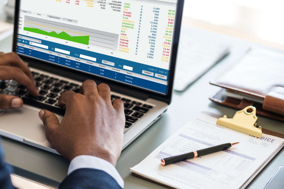 Pessoa utilizando um notebook com gráficos e dados financeiros exibidos na tela. Sobre a mesa há documentos, uma prancheta e uma caneta, representando análise financeira, investimentos, gestão de despesas e controle orçamentário