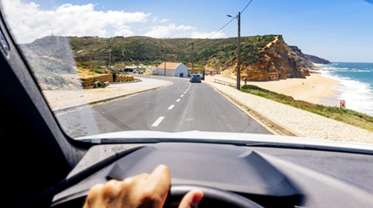 imagem de um painel de um carro, com a mão do cliente Itaú no volante e a vista da janela da frente de seu carro, uma estrada ao lado de uma praia.