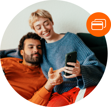 Um homem e uma mulher está sentado no sofá. Os dois estão sorrindo. A mulher segura um celular enquanto o homem aponta algo na tela do aparelho.