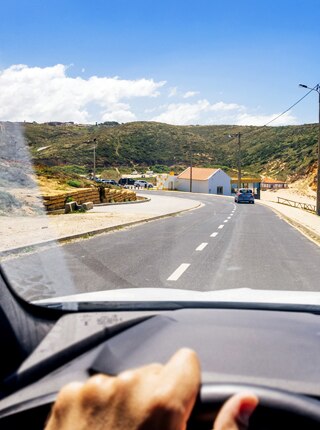 imagem de um painel de um carro, com a mão do cliente Itaú no volante e a vista da janela da frente de seu carro, uma estrada ao lado de uma praia.
