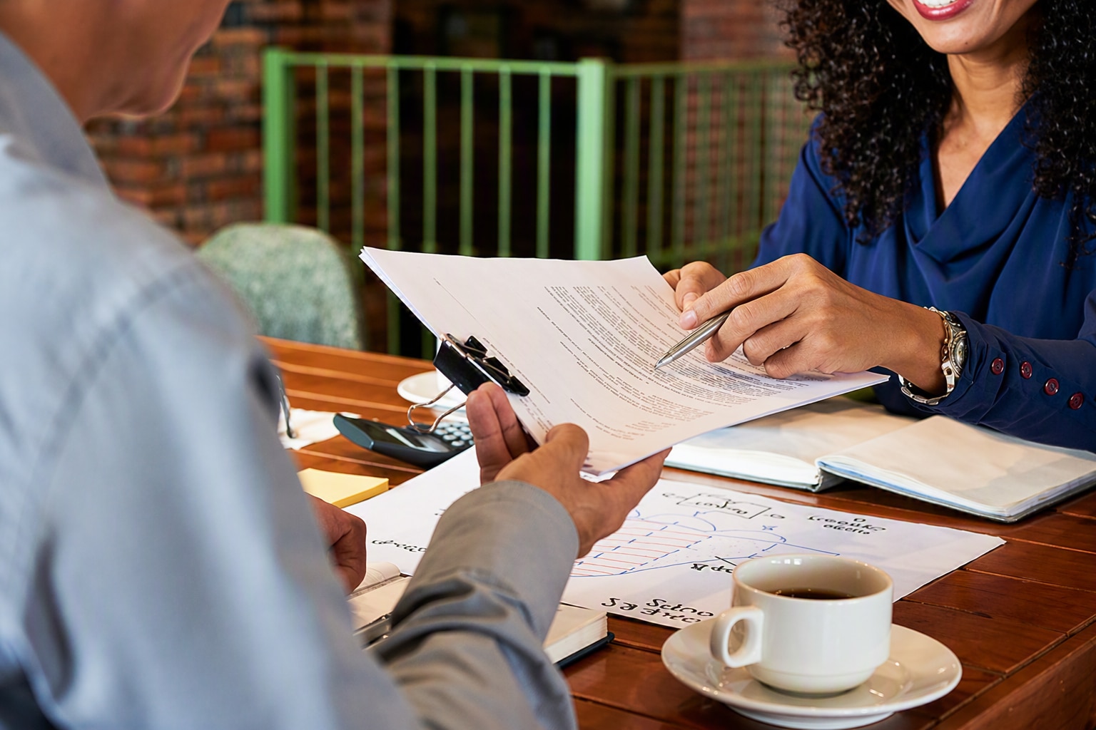 Pessoa em reunião de negócios analisando documentos sobre uma mesa, enquanto outra pessoa aponta informações em uma folha. Ao fundo, há materiais de trabalho e uma xícara de café.