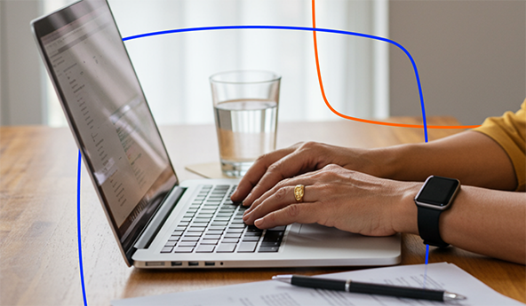 Pessoa digitando em um notebook em cima da mesa, com alguns objetos ao redor