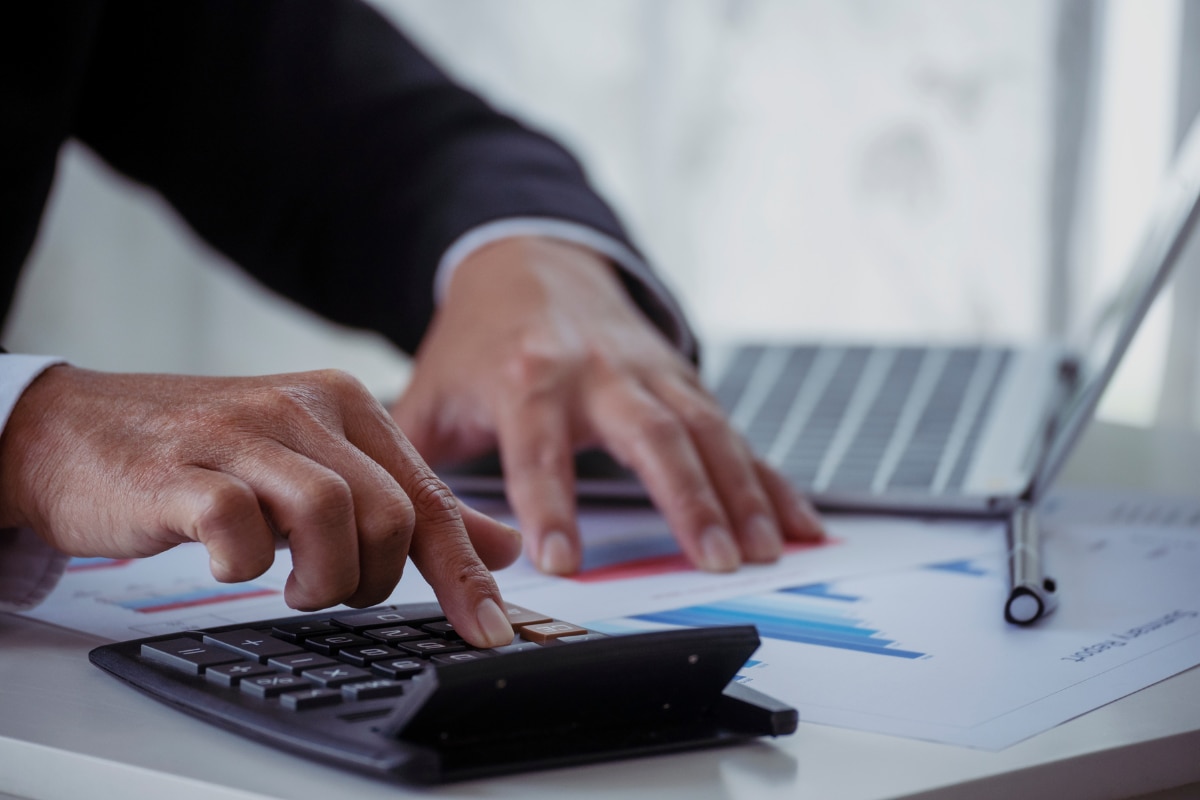 Pessoa utilizando uma calculadora sobre a mesa enquanto analisa relatórios financeiros com gráficos impressos. Ao fundo, há um notebook aberto apoiando a análise de dados e o planejamento financeiro da empresa.