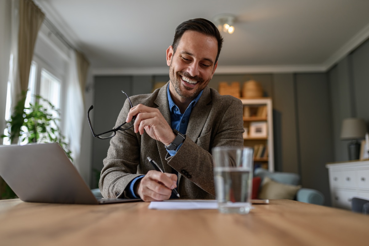 Copilot said: Pessoa trabalhando em uma mesa com notebook, escrevendo em uma folha de papel e segurando óculos, em ambiente interno.