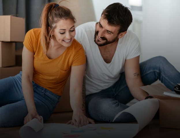 Um jovem casal olha para o projeto de reforma da casa. Ela é loira, pele clara e usa camieta laranja. Ele é moreno, pele branca, e usa camiseta branca. Ao fundo vemos caixas de mudança.