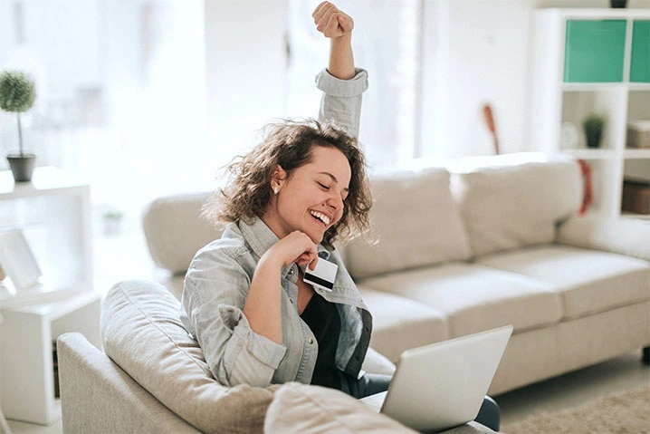 Mulher sentada no sofá da sua sala, sorridente, com seu notebook em seu colo, enquanto ergue uma mão para cima e com a outra, ela segura um cartão Itaú.