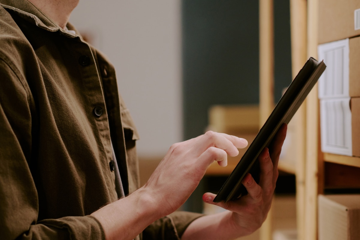 Pessoa utilizando um tablet preto em um ambiente de armazenamento, próxima a prateleiras com caixas organizadas. A mão toca a tela do dispositivo enquanto a outra o segura. A cena tem iluminação interna suave e tons quentes, sugerindo atividades de controle de estoque, logística ou inventário.