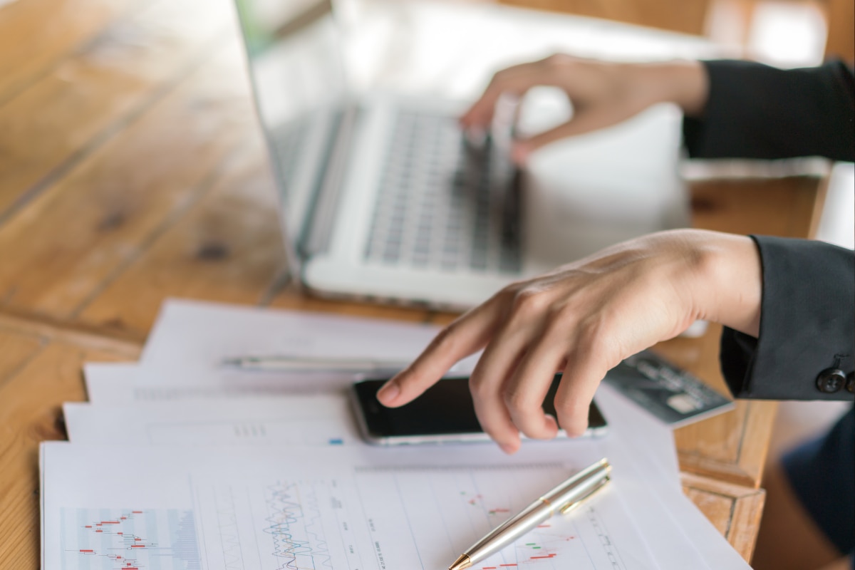 Mãos utilizando smartphone e notebook sobre mesa com relatórios financeiros e gráficos, representando gestão financeira e planejamento empresarial.