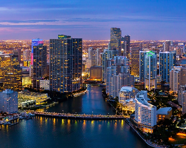 Imagem de um centro urbano a noite com prédios iluminados e uma ponte.