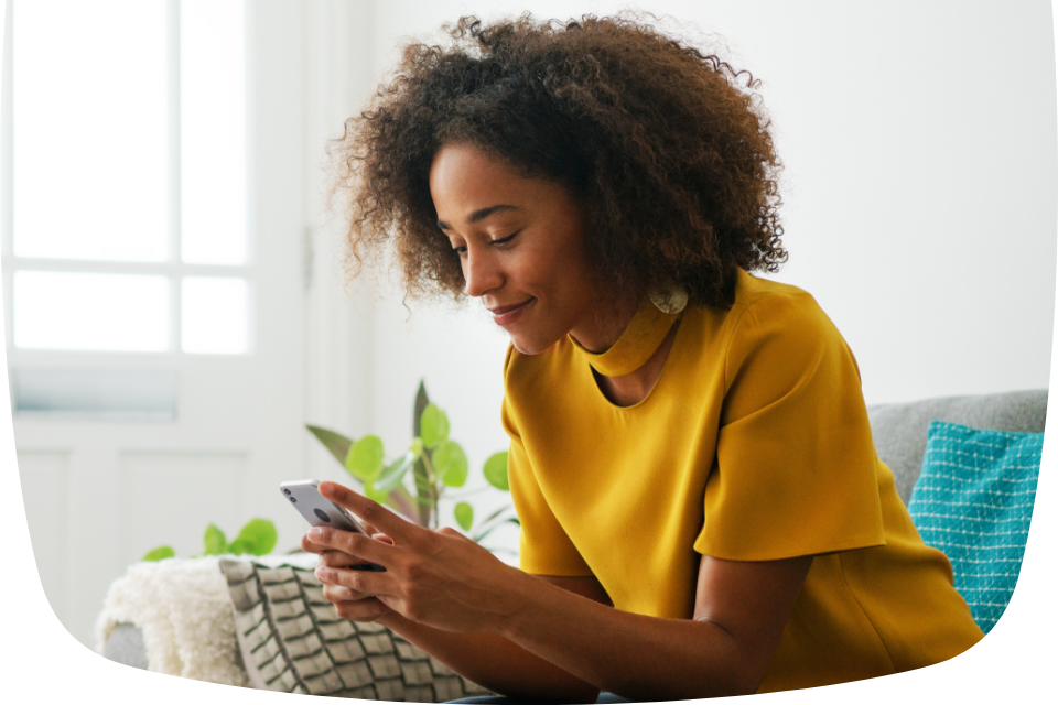 Mulher negra com blusa amarela sentada em um sofá, mexendo no celular e sorrindo levemente.
