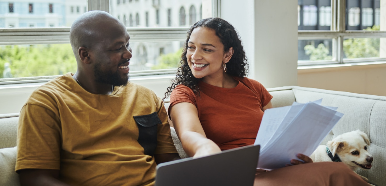 casal sentado no sofá com um cachorrinho, apontando para a tela de um notebook com sorriso no rosto.