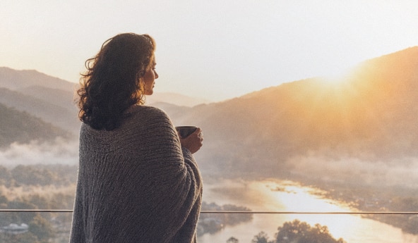 Pessoa observa paisagem montanhosa ao amanhecer segurando uma caneca.
