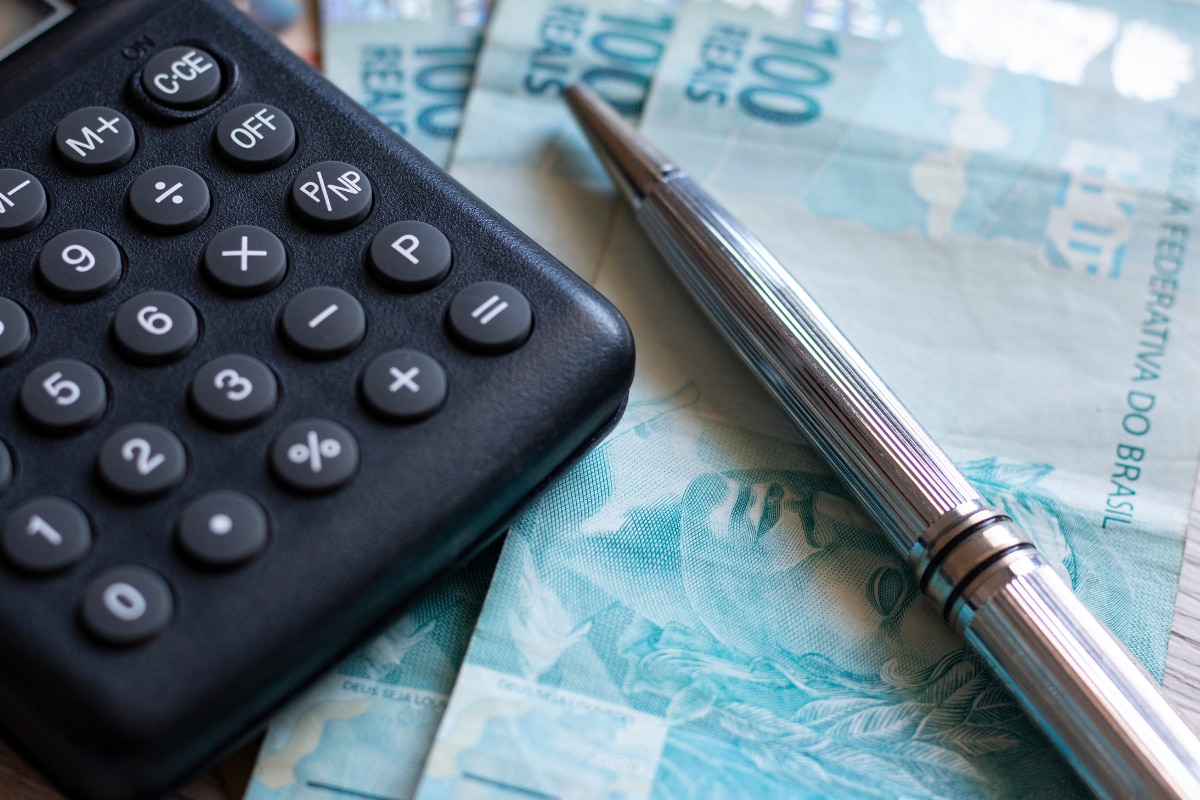 Calculadora preta e caneta metálica prateada sobre cédulas de 100 reais, representando cálculos financeiros, orçamento, despesas e planejamento econômico.