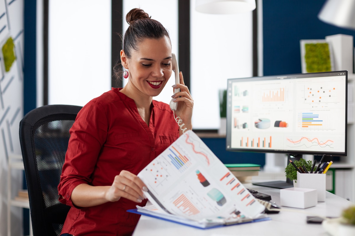 Pessoa em escritório utilizando telefone de mesa enquanto analisa relatórios impressos. Ao lado, monitor exibe gráficos e indicadores de desempenho em uma estação de trabalho organizada.