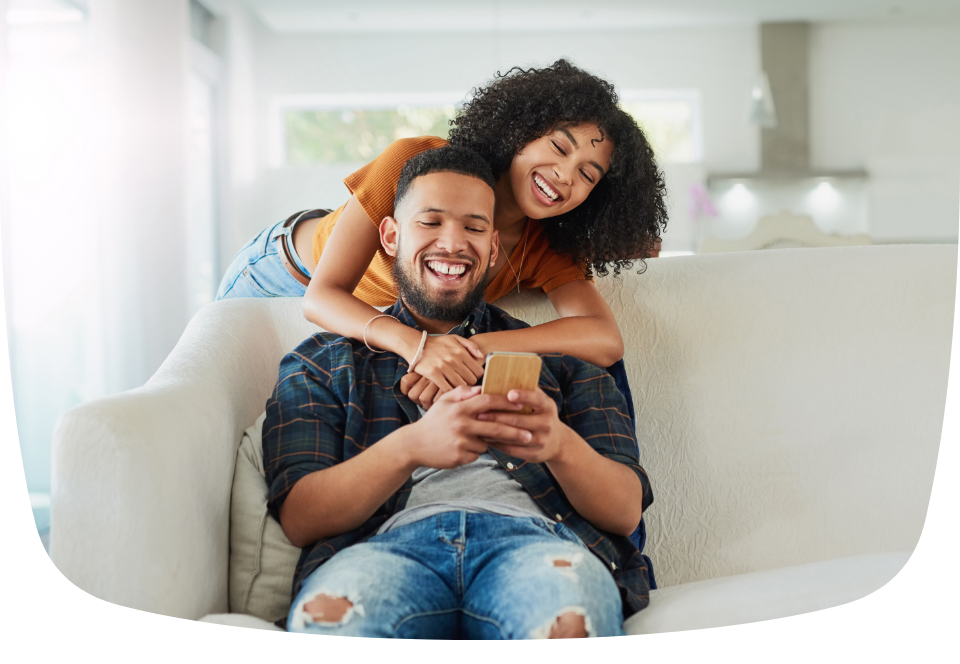 Um homem e uma mulher está sentado no sofá. Os dois estão sorrindo. A mulher segura um celular e o homem está abraçado com ela.