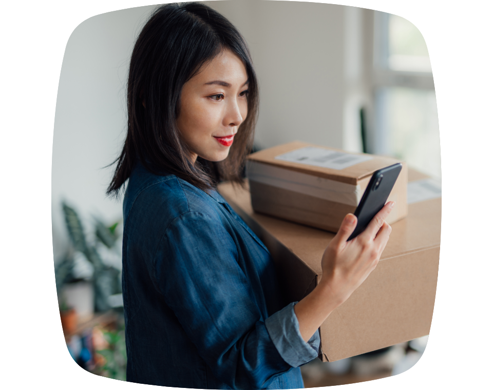 Mulher com camisa jeans segurando algumas caixas em uma das mãos e olhando para o celular.