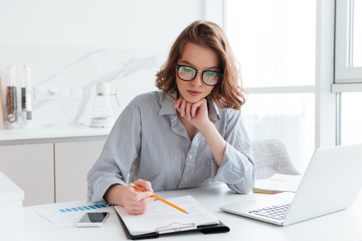 Pessoa utilizando óculos sentada em uma mesa, analisando documentos em uma prancheta enquanto faz anotações com um lápis. Sobre a mesa há um notebook aberto, um celular e folhas com informações impressas, em um ambiente de trabalho iluminado.